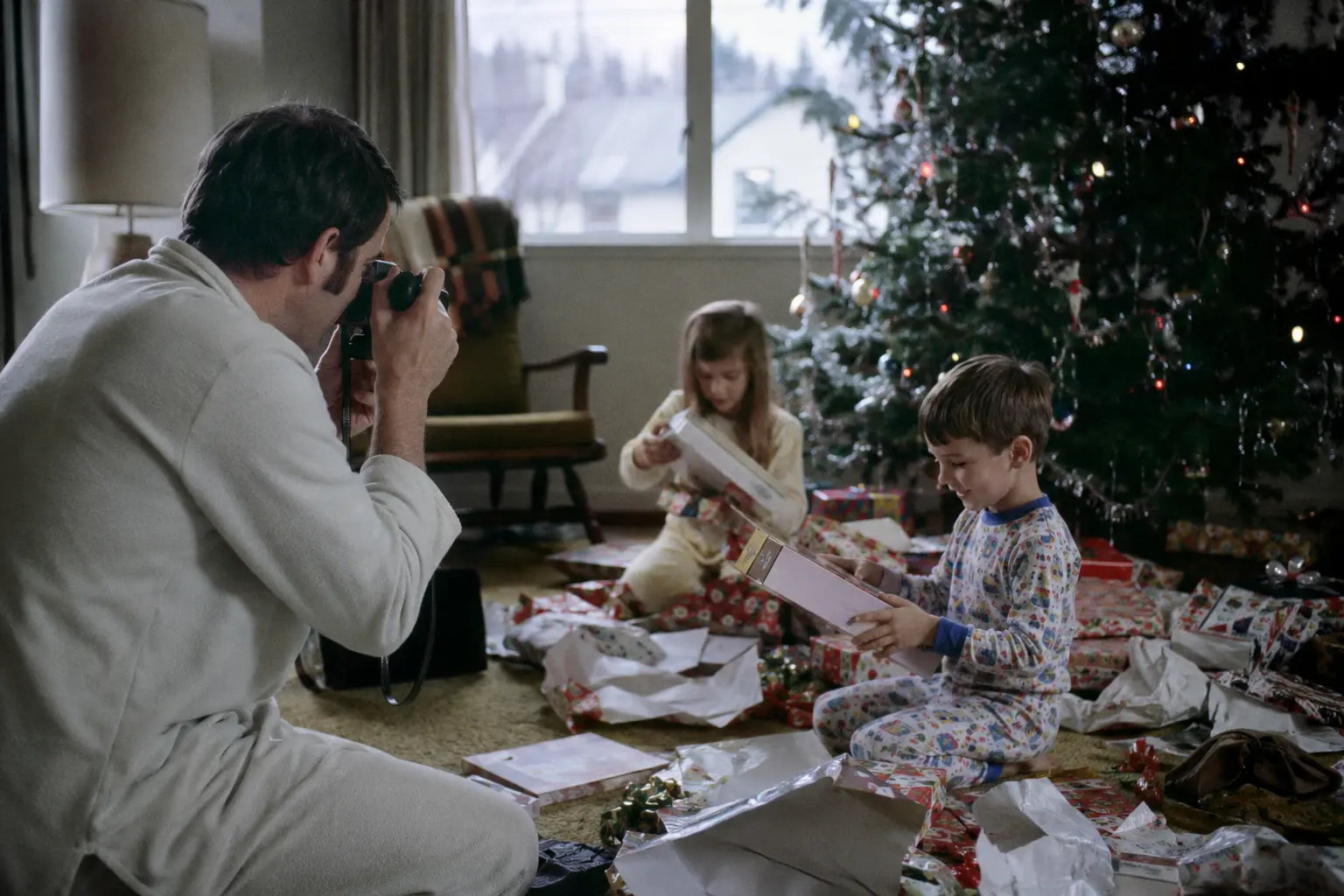 A parent photographing children on Christmas morning with a film camera in the 1970s
